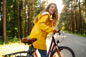 Smiling woman with curly hair in a yellow coat rides a bicycle in a sunny park.